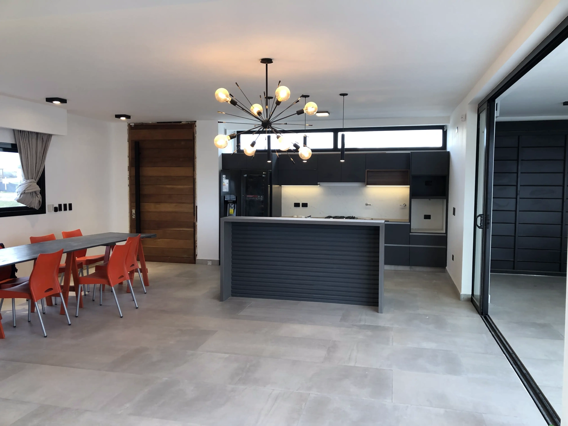 Foto de una casa en puerto del Águila construída con el sistema de Steel Framing por Vértice Construcciones en la que se observa una sala de estar con una cocina con una barra en forma de isla y un comedor con mesa y sillas en el interior de la casa y la puerta principal al fondo
