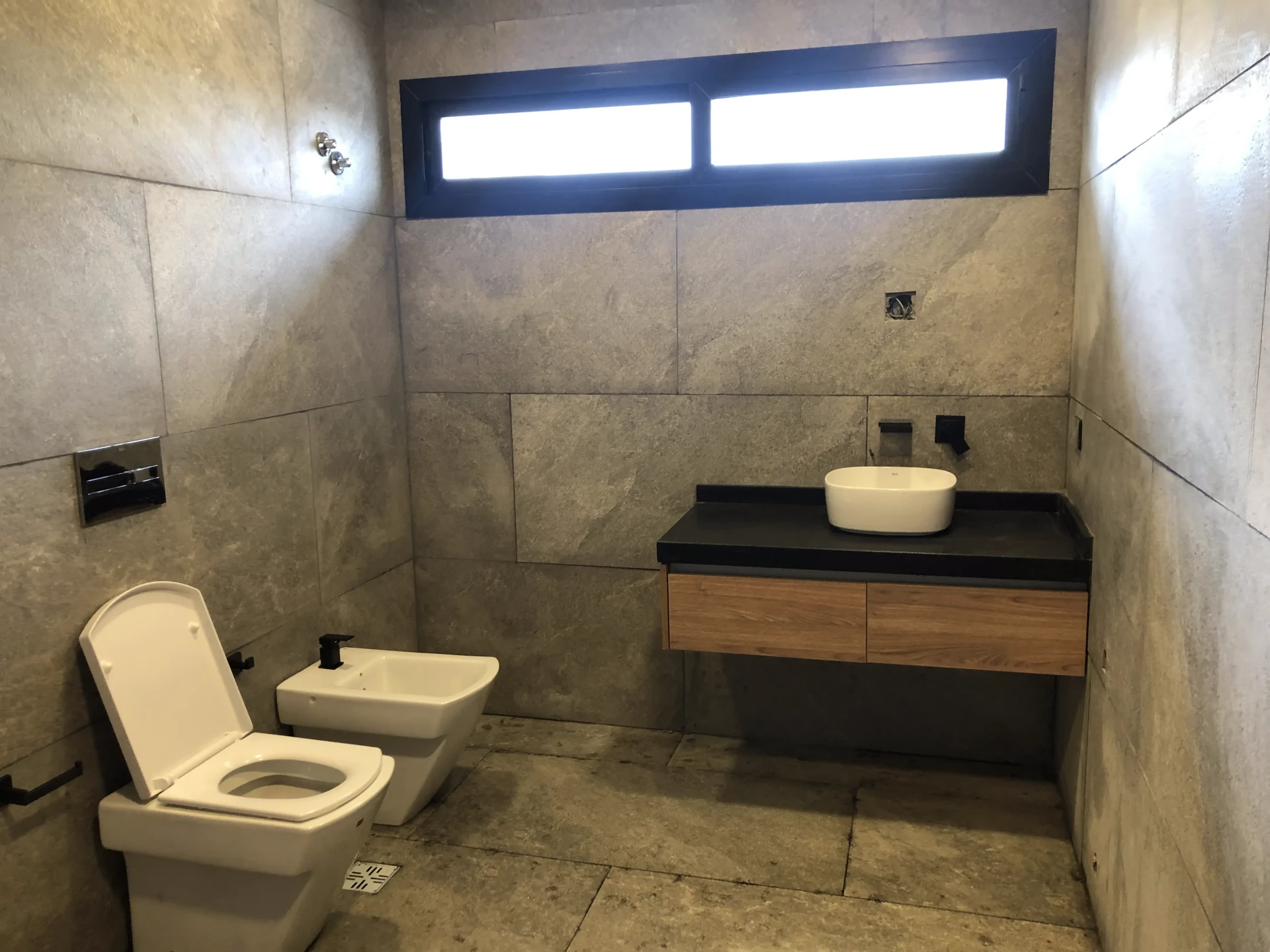 Foto de una casa en puerto del Águila construída con el sistema de Steel Framing por Vértice Construcciones en la que se observa un baño con inodoro, bidet y mesada con una bacha para lavarse las manos, todas las paredes revestido con cerámico, y una de ellas con una ventana rectangular en la parte superior