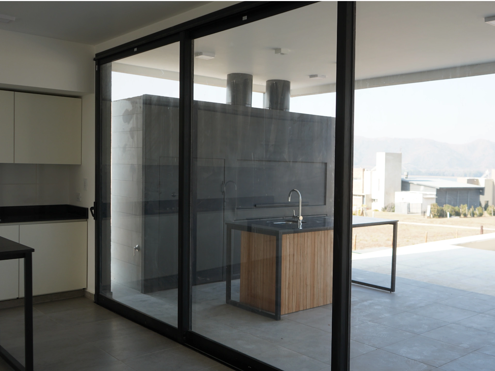Foto de una casa en puerto del Águila construída con el sistema de Steel Framing por Vértice Construcciones en la que se observa desde adentro de la casa, a través de los ventanales, la galería con la mesada tipo barra y el asador