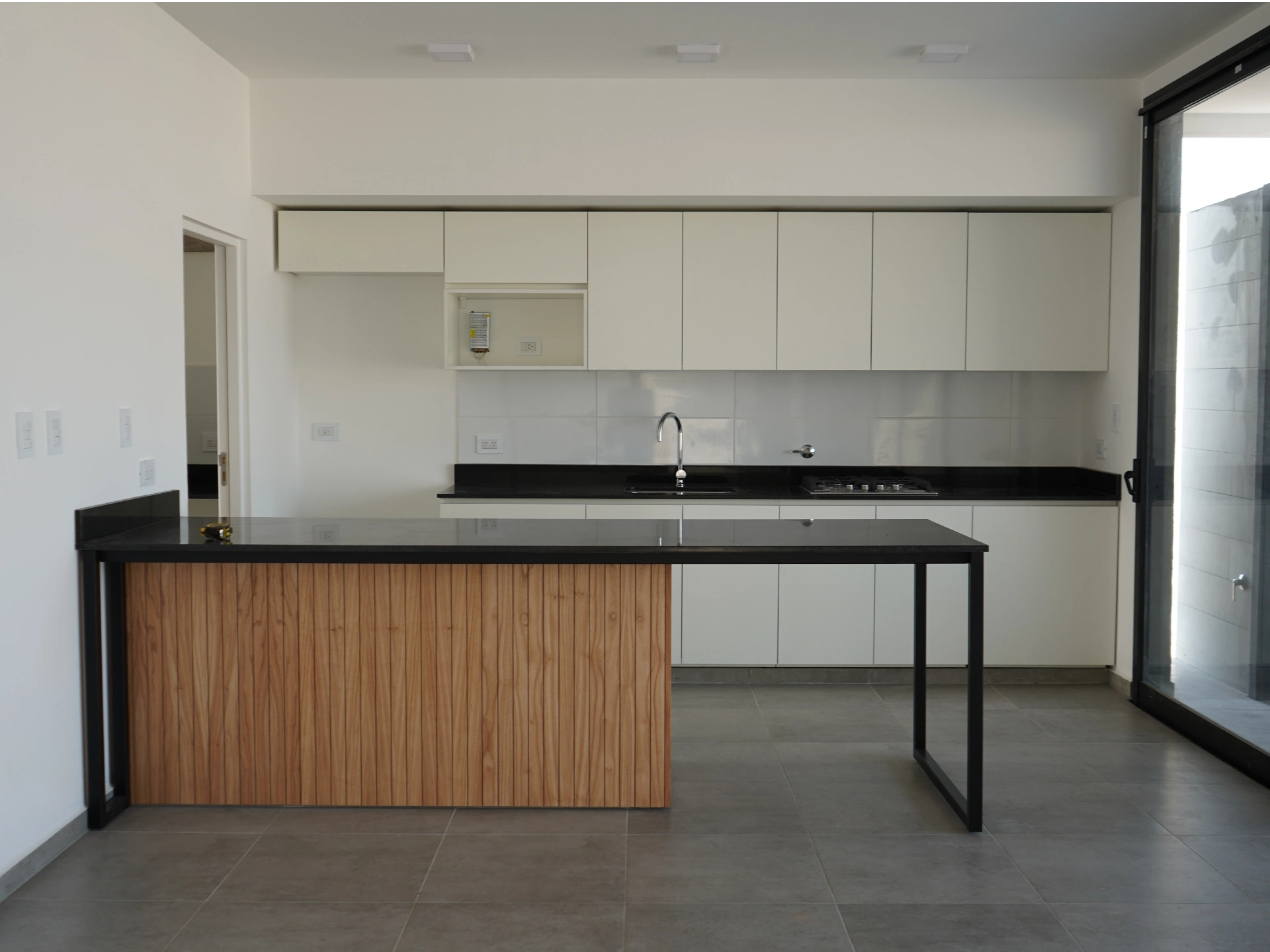 Foto de una casa en puerto del Águila construída con el sistema de Steel Framing por Vértice Construcciones en la que se observa en primer plano la cocina con muebles blancos y una mesada de mármol negro idéntica a la que tiene la mesada en forma de isla revestida con madera
