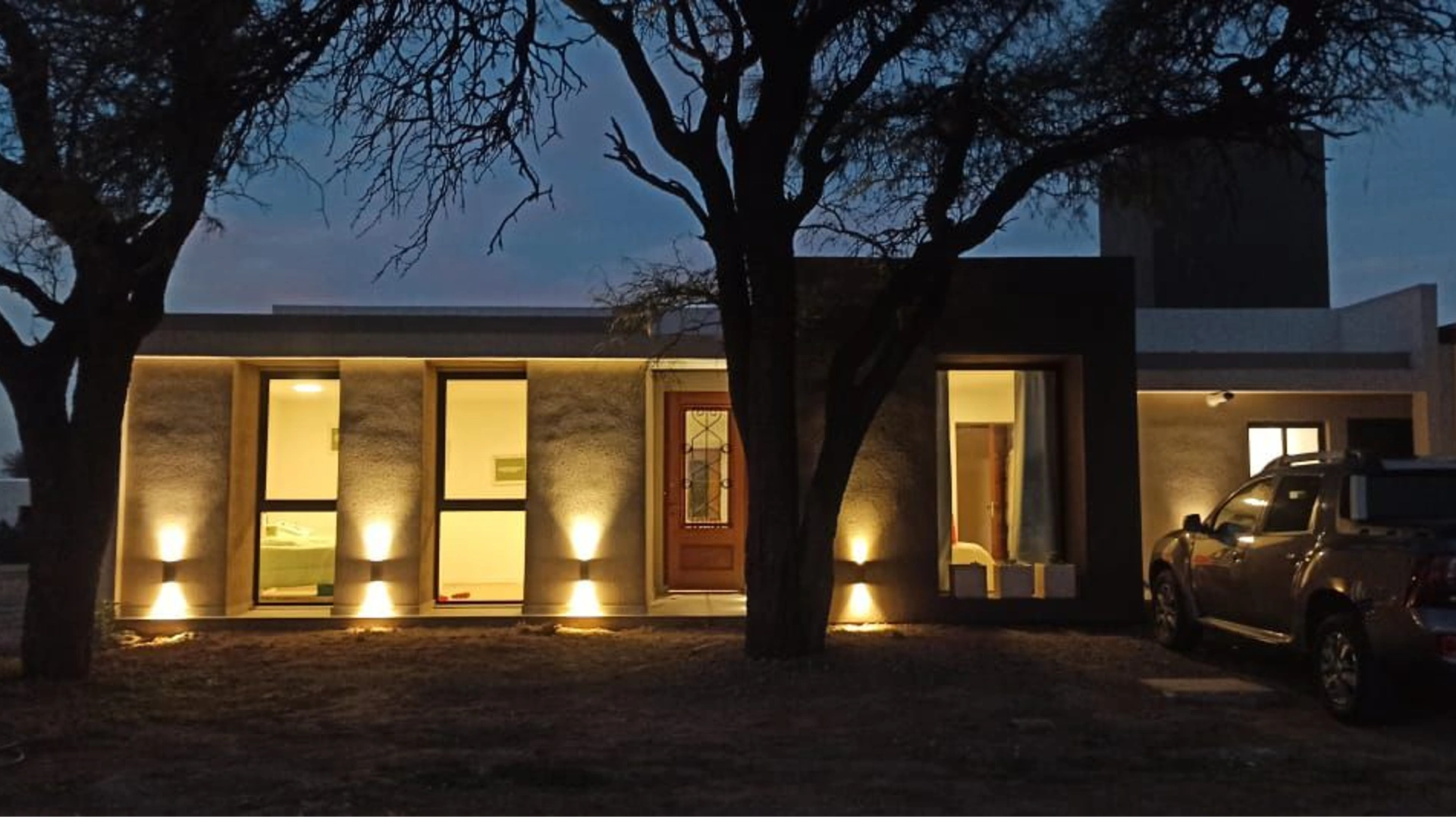 Foto de una casa en 7 Soles construída con el sistema de Steel Framing por Vértice Construcciones en la que se observa la fachada de la casa revestida con el sistema EIF´s, iluminada con luces prendidas ya que es de noche, varias ventanas de techo a piso y dos árboles en el frente, detrás de los cuales se encuentra la puerta de ingreso de madera con un vidrio en el centro detrás de unas rejas negras ornamentadas