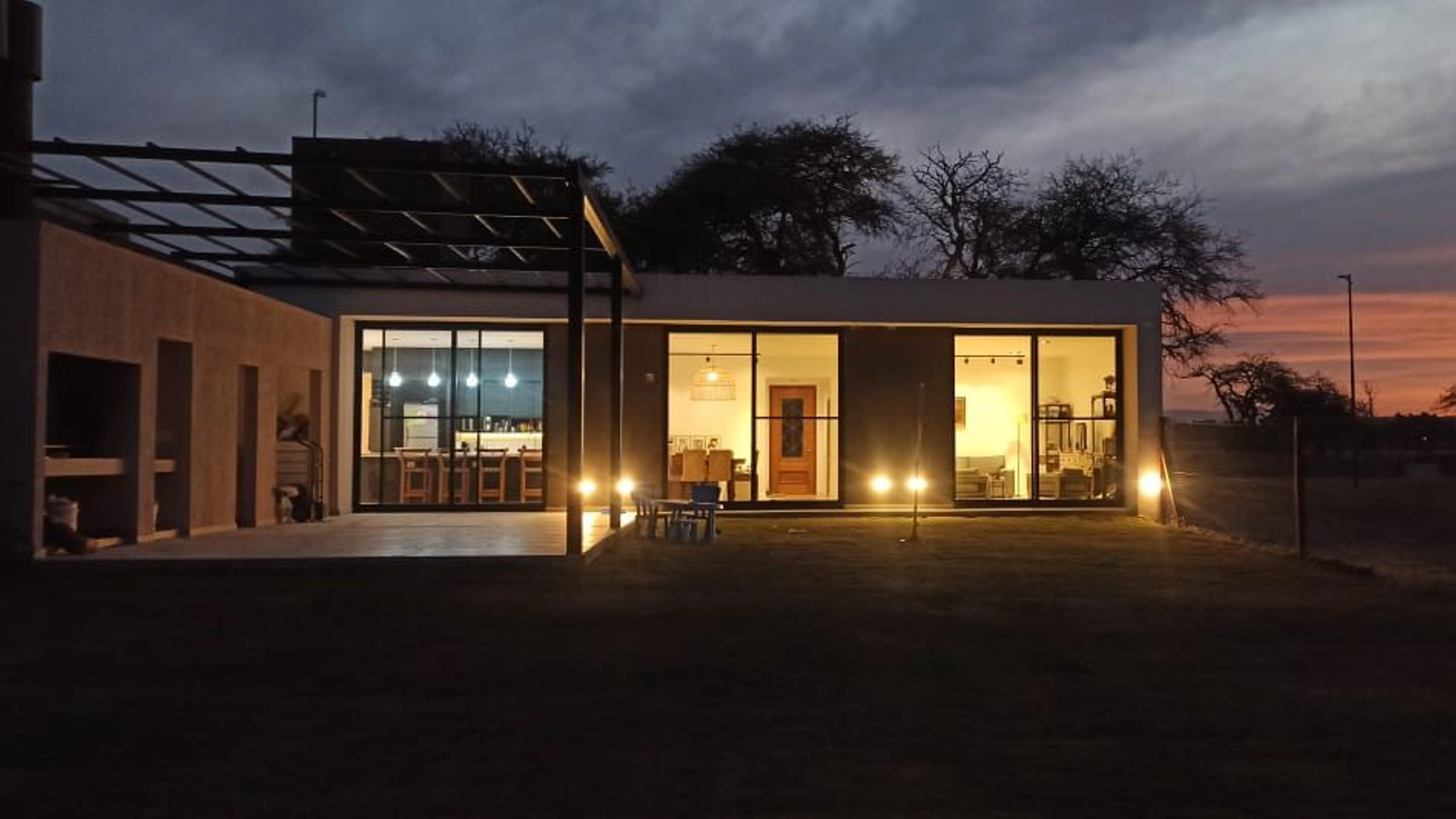 Foto de una casa en 7 Soles construída con el sistema de Steel Framing por Vértice Construcciones en la que se observa el patio con césped y más atrás la galería con una pérgola de hierro negro y la fachada trasera de la casa revestida con el sistema EIF´s, iluminada con luces prendidas ya que es de noche, con tres ventanales grandes de techo a piso a través de los cuales se observa una sala de estar interior