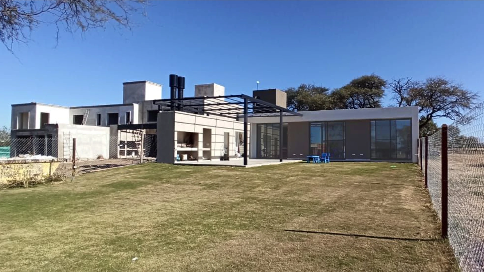 Foto de una casa en 7 Soles construída con el sistema de Steel Framing por Vértice Construcciones en la que se observa el patio con césped y más atrás la galería con una pérgola de hierro negro y un asador y la fachada trasera de la casa revestida con el sistema EIF´s, con tres ventanales grandes de techo a piso y que se ven espejados ya que es de día y hay mucha luz por el sol
