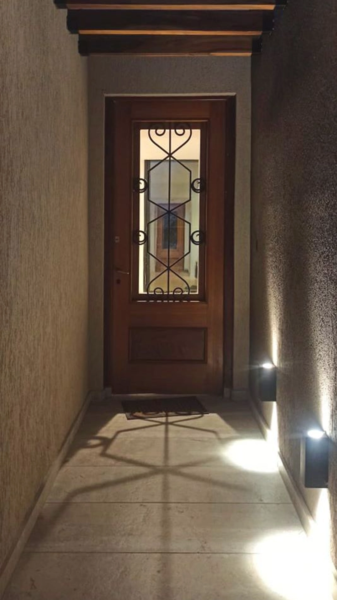 Foto de una casa en 7 Soles construída con el sistema de Steel Framing por Vértice Construcciones en la que se observa en primer plano el pasillo de ingreso iluminado con luces prendidas ya que es de noche y al fondo una puerta de ingreso de la casa de madera con un vidrio en el centro detrás de unas rejas negras ornamentadas