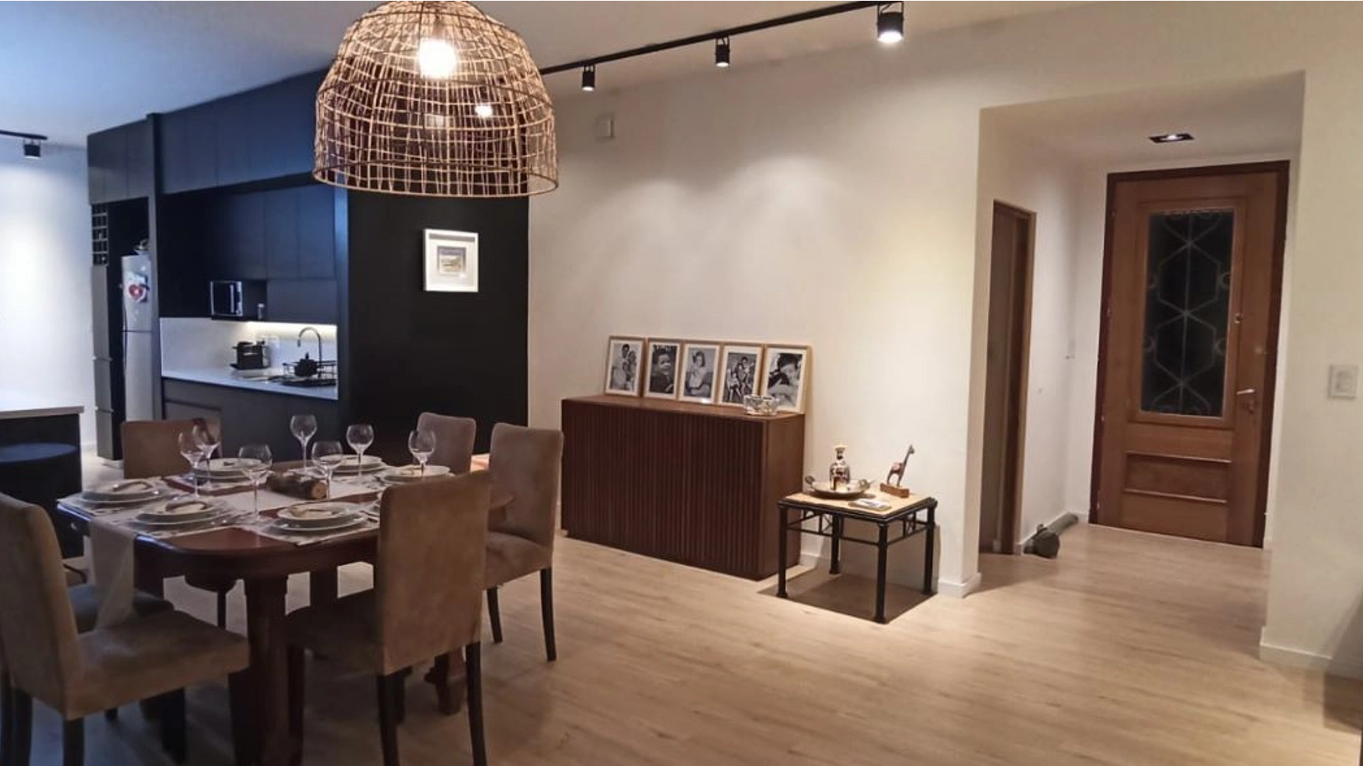 Foto de una casa en 7 Soles construída con el sistema de Steel Framing por Vértice Construcciones en la que se observa la sala de estar interior con una mesa puesta y sillas con una lámpara de yute colgada del techo y detrás un mueble de madera con varios portarretratos con fotos de una familia, hacia un costado la cocina con muebles de madera marrón oscuro y hacia otro lado se ve la puerta de ingreso de madera con un vidrio en el centro
