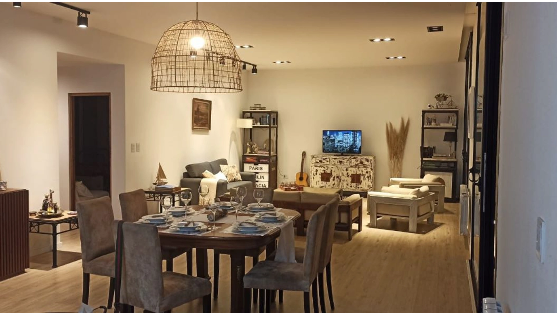 Foto de una casa en 7 Soles construída con el sistema de Steel Framing por Vértice Construcciones en la que se observa la sala de estar interior con una mesa puesta y sillas con una lámpara de yute colgada del techo y detrás un juego de sillones, un televisor sobre un mueble entre dos estanterías de hierro con varios objetos