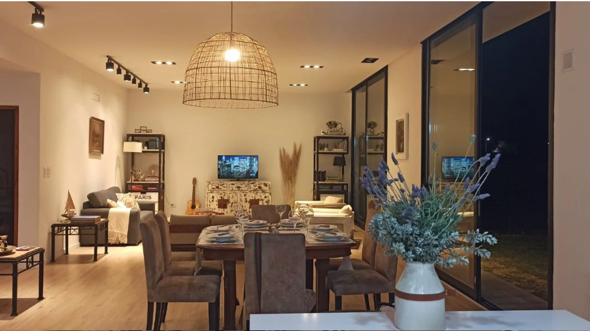 Foto de una casa en 7 Soles construída con el sistema de Steel Framing por Vértice Construcciones en la que se observa la sala de estar interior con una mesa puesta y sillas con una lámpara de yute colgada del techo y detrás un juego de sillones, un televisor sobre un mueble entre dos estanterías de hierro con varios objetos. De lado derecho de la foto se ven dos grandes ventanaes que dan hacia un patio externo