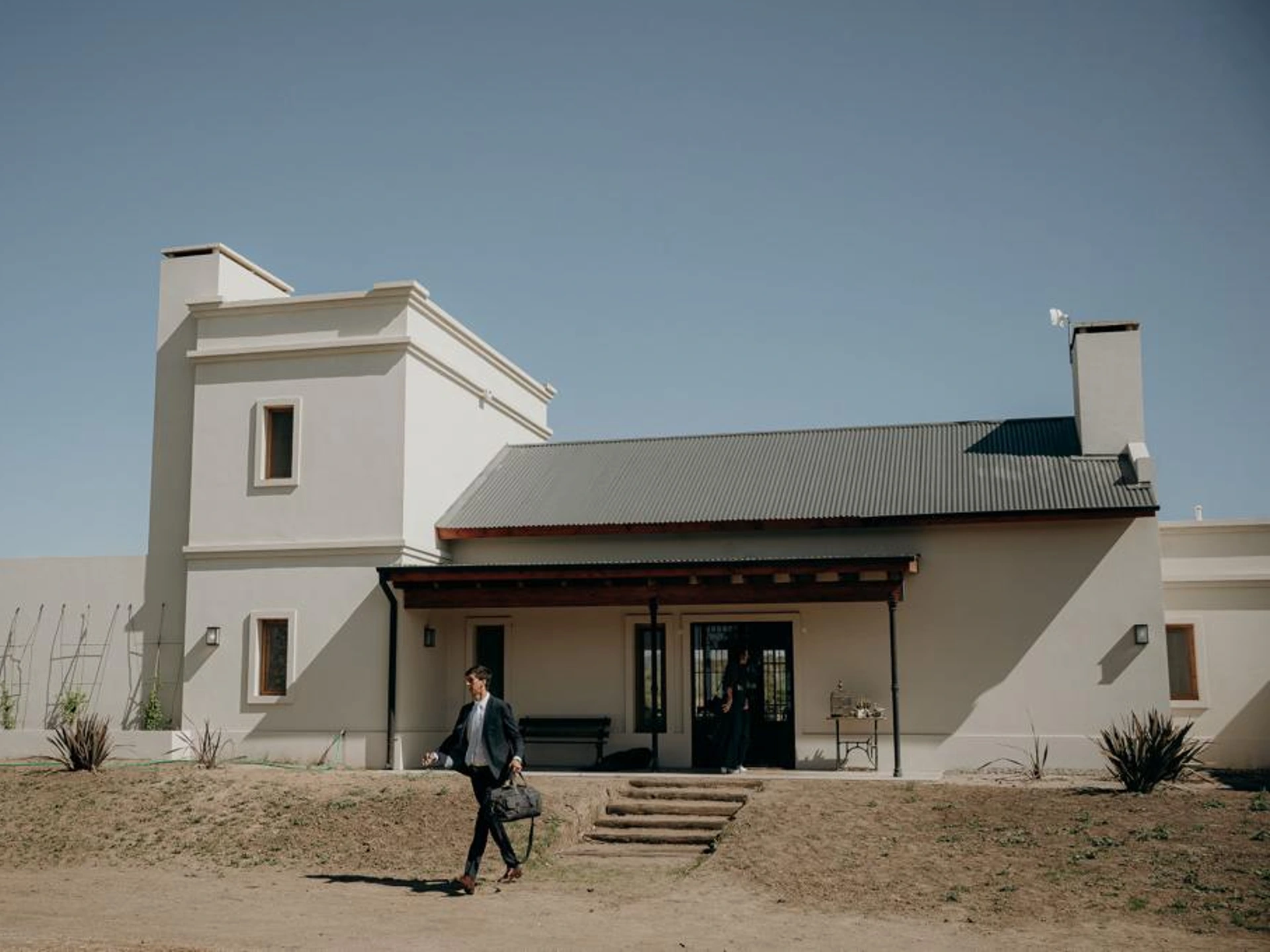Foto de una casa en Ascochinga construída con el sistema tradicional por Vértice Construcciones en la que se observa la fachada de la casa de estilo colonial pintada de blanco mate con aberturas de madera, techos de chapa a dos aguas y una galería pequeña al frente donde se ubica la puerta de ingreso de hierro negro y vidrio; bajando las escaleras del ingreso de la casa hay un hombre alto de traje