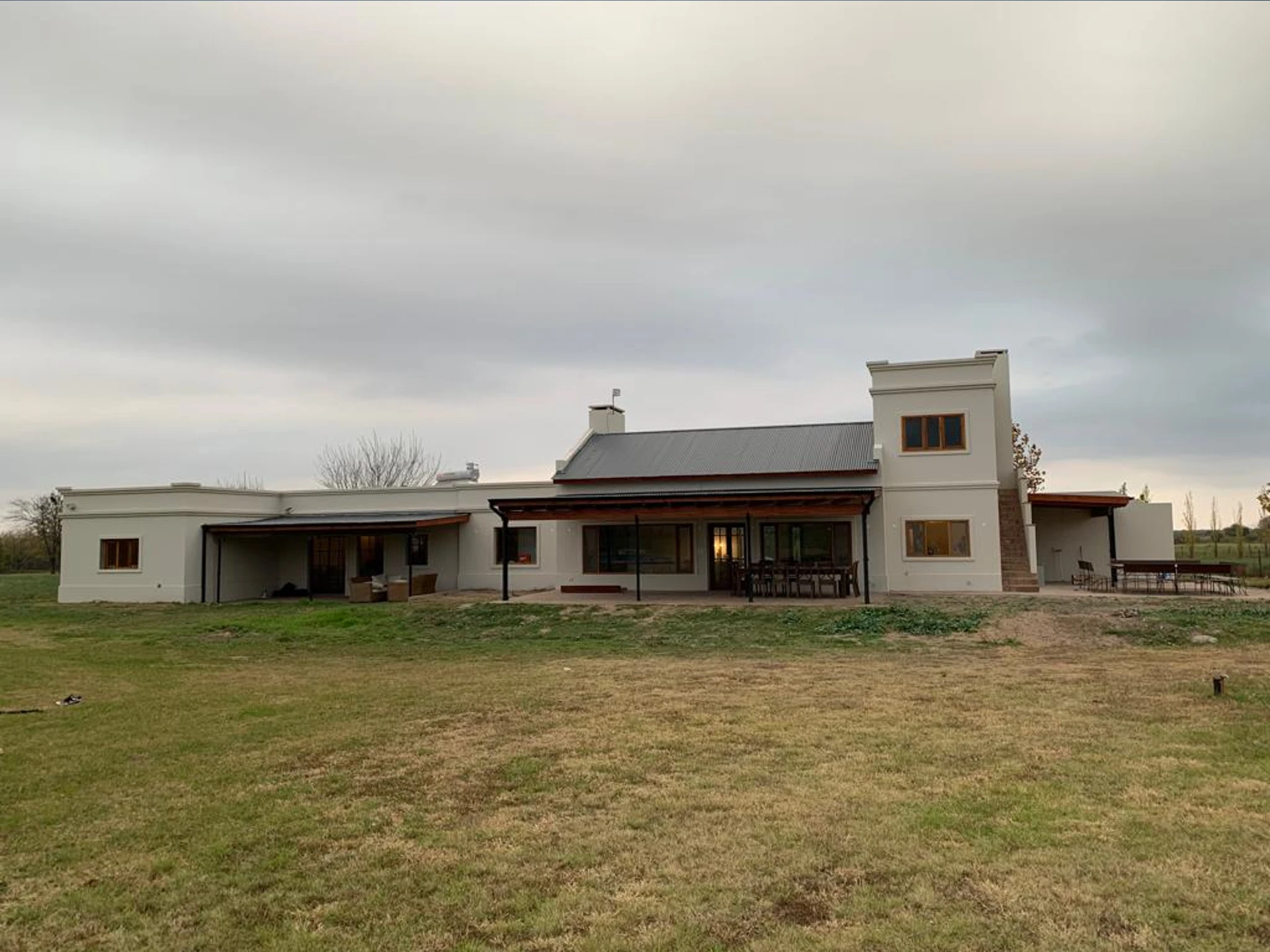 Foto de una casa en Ascochinga construída con el sistema tradicional por Vértice Construcciones en la que se observa la fachada trasera de la casa de estilo colonial pintada de blanco mate con aberturas de madera, techos de chapa a dos aguas y tres galerías con techo de chapa, ubicadas de manera independiente a lo largo de la casa; dos de esas galerías tienen mesas y sillas