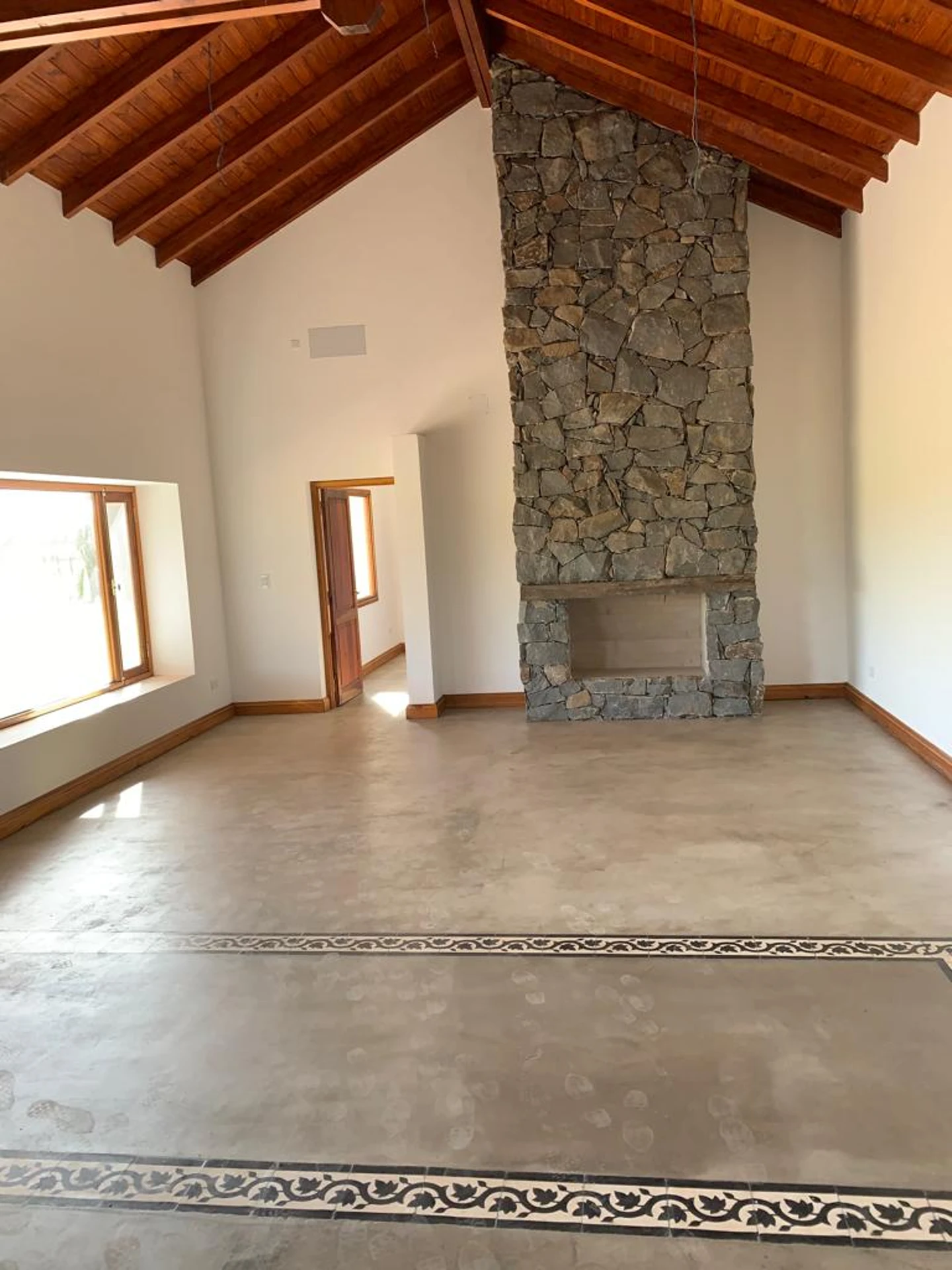 Foto de una casa en Ascochinga construída con el sistema tradicional por Vértice Construcciones en la que se observa una sala de estar interior vacía con un piso de microcemento y algunos cerámicos de estilo colonial, un techo alto revestido de madera y una gran chimenea revestida de piedra, hacia un costado hay una gran ventana con abertura de madera y alfeizar, por la que entra mucha luz