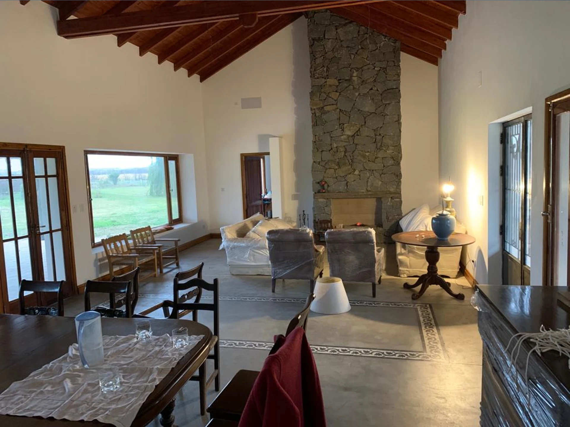 Foto de una casa en Ascochinga construída con el sistema tradicional por Vértice Construcciones en la que se observa una sala de estar interior en plena mudanza con un piso de microcemento y algunos cerámicos de estilo colonial, un techo alto revestido de madera y una gran chimenea revestida de piedra, hacia un costado hay una gran ventana con abertura de madera y alfeizar; también hay algunos muebles con y sin cinta de embalar, entre los cuales se destaca una mesa, sillas y sillones