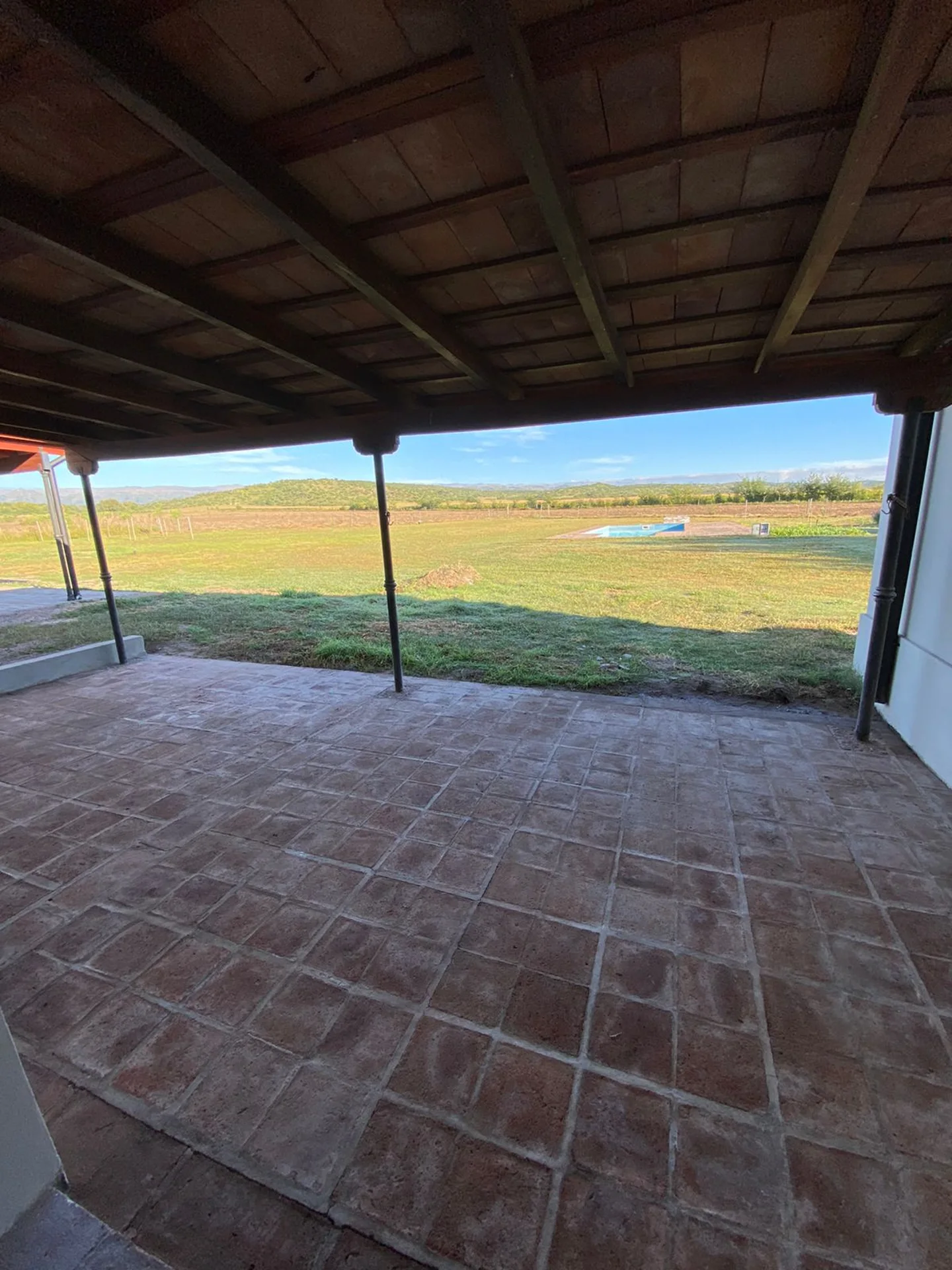 Foto de una casa en Ascochinga construída con el sistema tradicional por Vértice Construcciones en la que se observa una galería exterior con piso de ladrillo y techo revestido de madera, y al fondo, un amplio jardín y una pileta, sin otras edificaciones a la vista