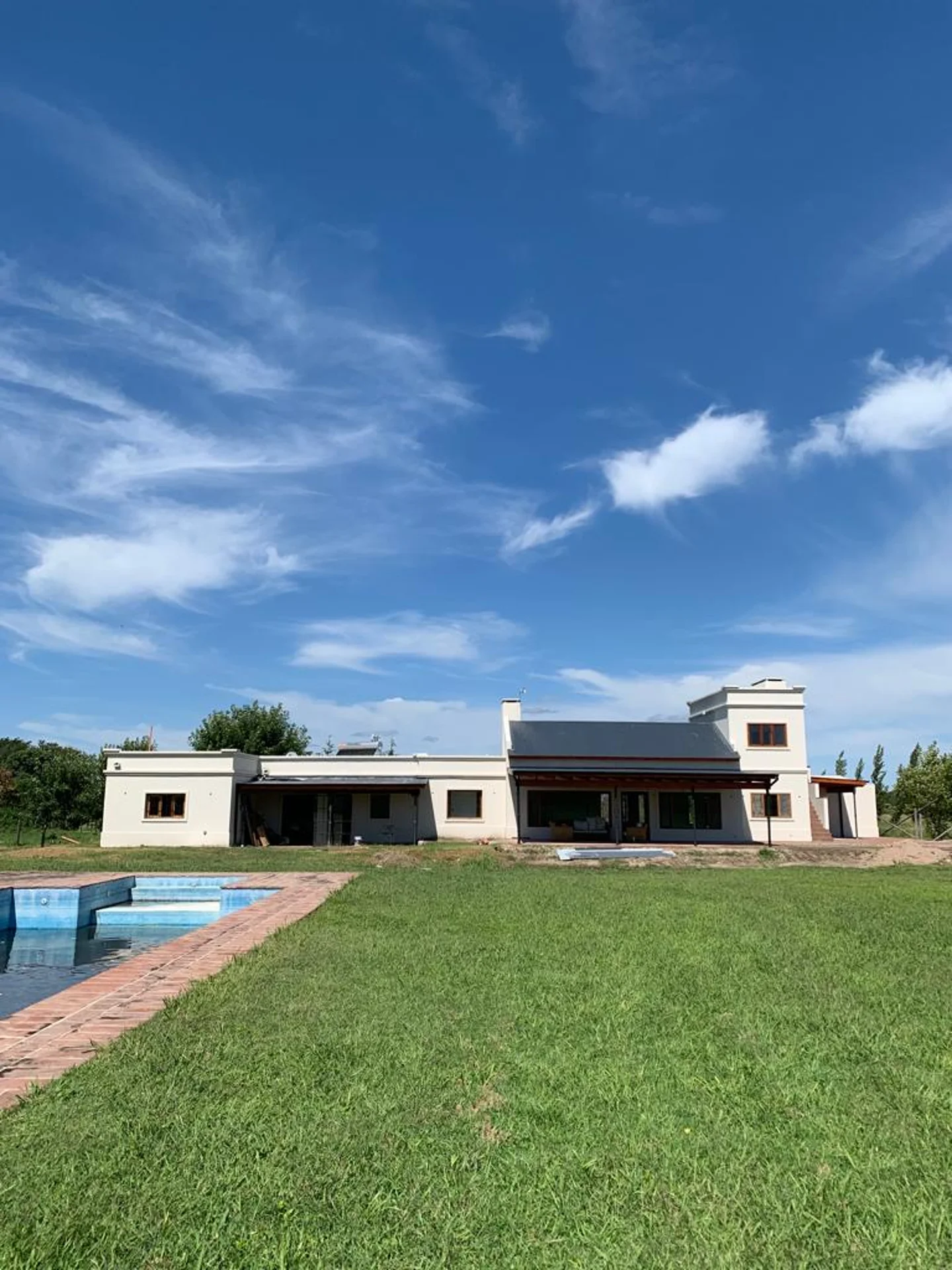 Foto de una casa en Ascochinga construída con el sistema tradicional por Vértice Construcciones en la que se observa en primer plano parte de una pileta con piso de ladrillo  y al fondo la fachada trasera de la casa con estilo colonial pintada de blanco mate con aberturas de madera, techos de chapa a dos aguas y tres galerías con techo de chapa, ubicadas de manera independiente a lo largo de la casa
