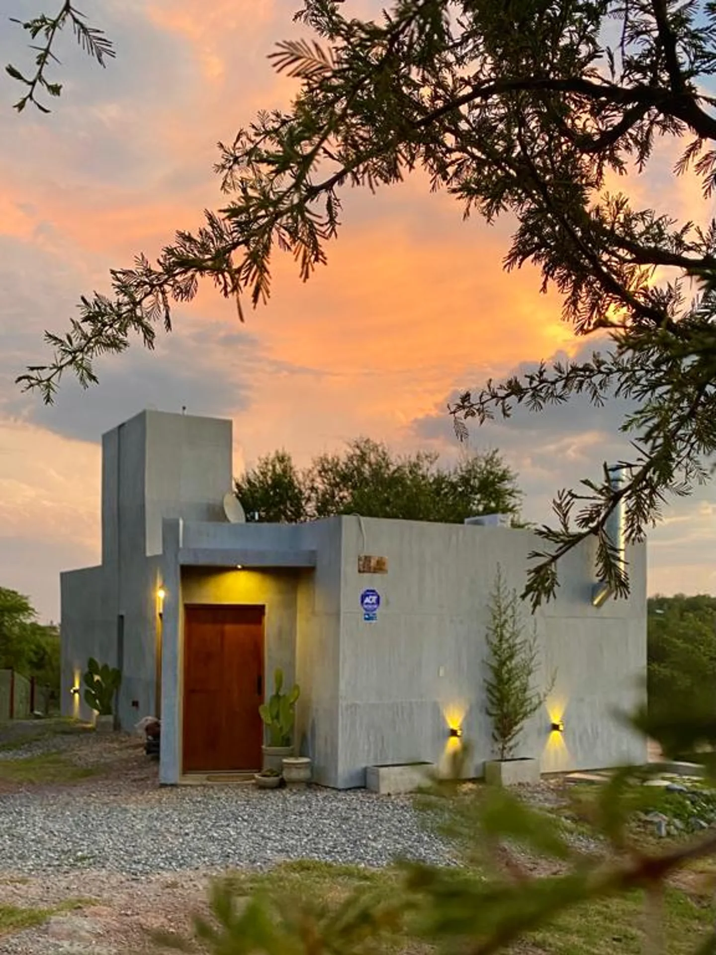 Foto de una casa en Bialet Massé construída con el sistema de Steel Framing por Vértice Construcciones en la que se observa la fachada principal de la casa con un acabado símil hormigón visto y una puerta principal de madera lisa, y varias macetas de hormigón con plantas de clima seco como cactus y espinillos