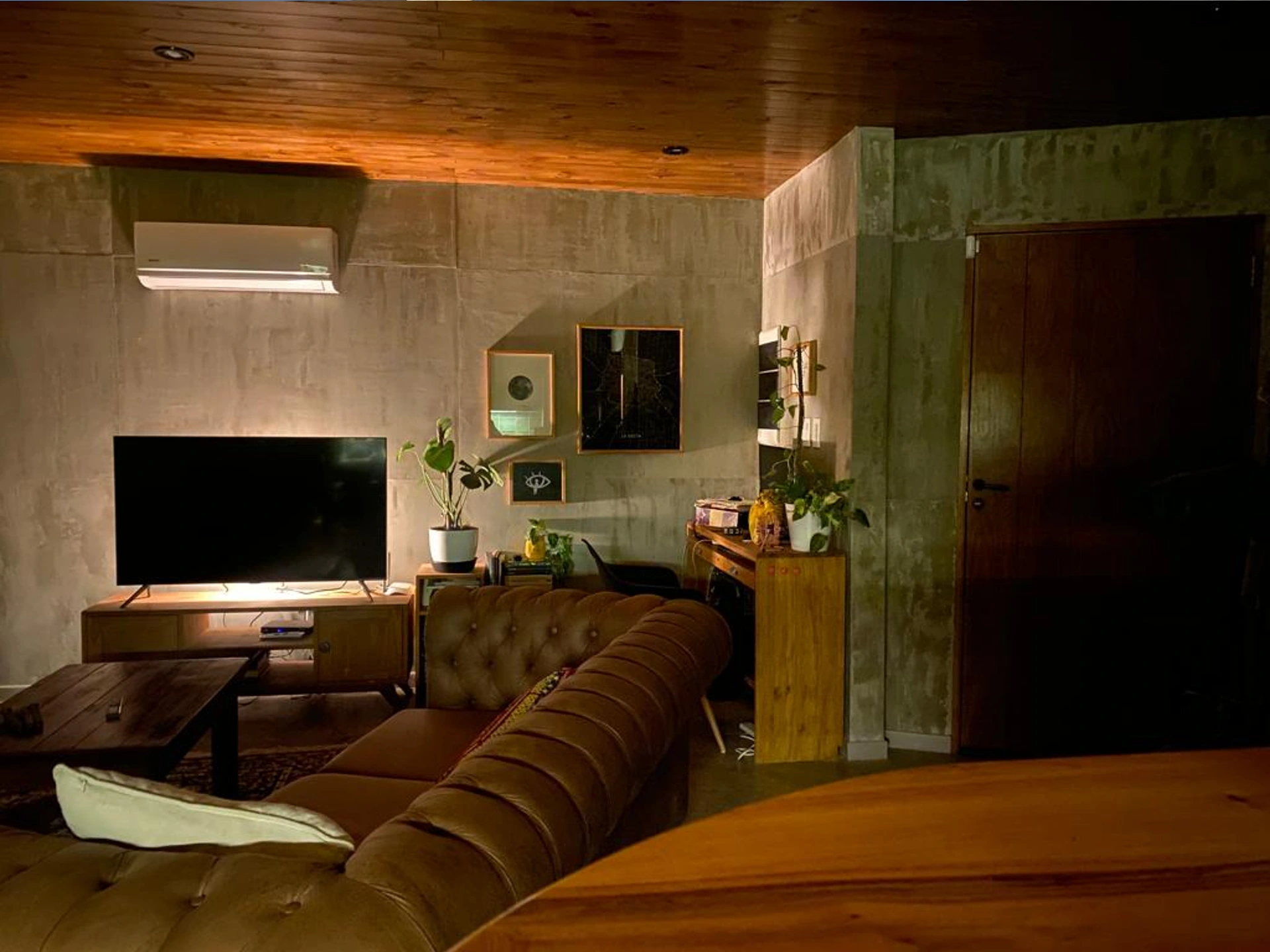 Foto de una casa en Bialet Massé construída con el sistema de Steel Framing por Vértice Construcciones en la que se observa una sala de estar interior con muebles de madera y un sillón grande de símil cuero; el piso es de hormigón alisado color gris plomo, el techo de madera y hay varias macetas con plantas de clima seco