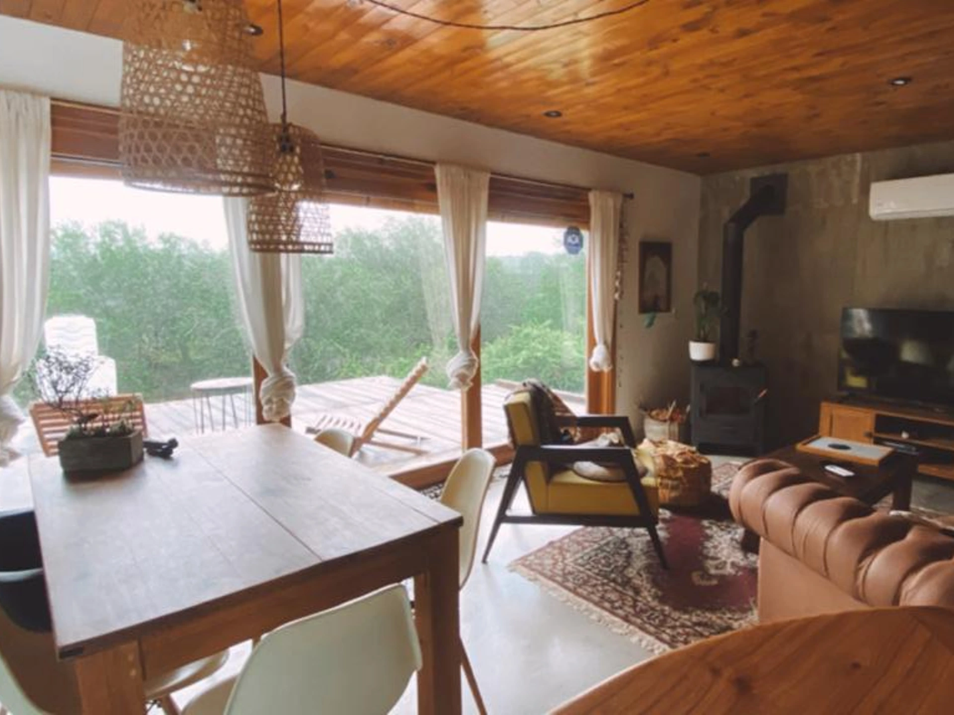 Foto de una casa en Bialet Massé construída con el sistema de Steel Framing por Vértice Construcciones en la que se observa una sala de estar interior con un sector de sillones y muebles de madera con una alfombra y otro sector con una mesa de madera y sillas tipo comedor y dos lámparas colgantes de mimbre; detrás hay un gran ventanal a través del cual se observa un deck de madera con muebles de exterior