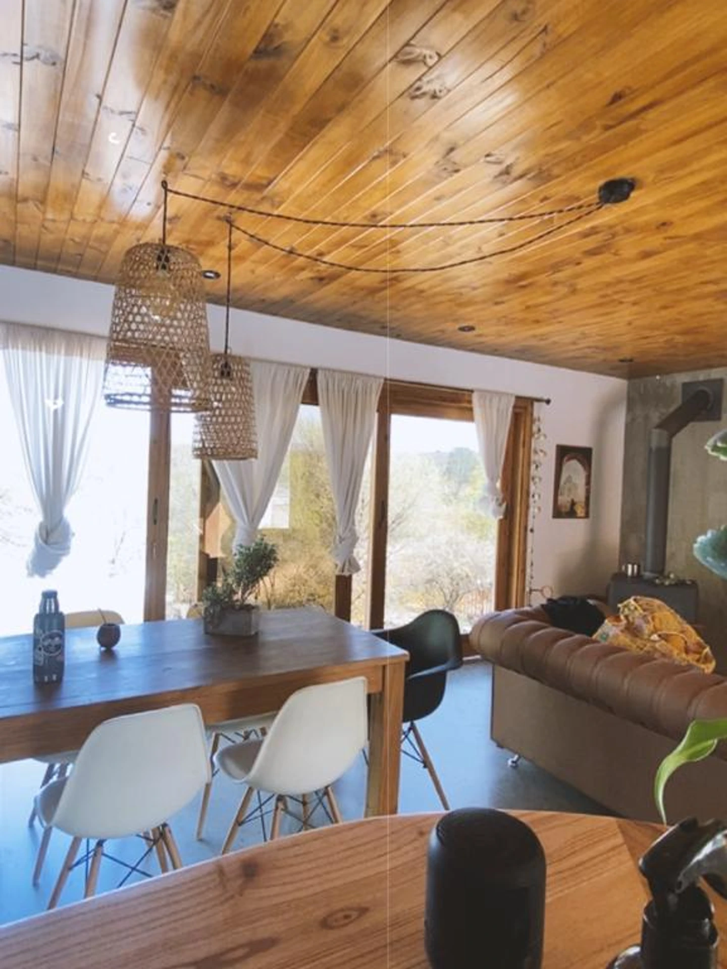Foto de una casa en Bialet Massé construída con el sistema de Steel Framing por Vértice Construcciones en la que se observa una sala de estar interior con un sector de sillones y muebles de madera y otro sector con una mesa de madera y sillas tipo comedor y dos lámparas colgantes de mimbre; detrás hay un gran ventanal con vista a las sierras de Córdoba