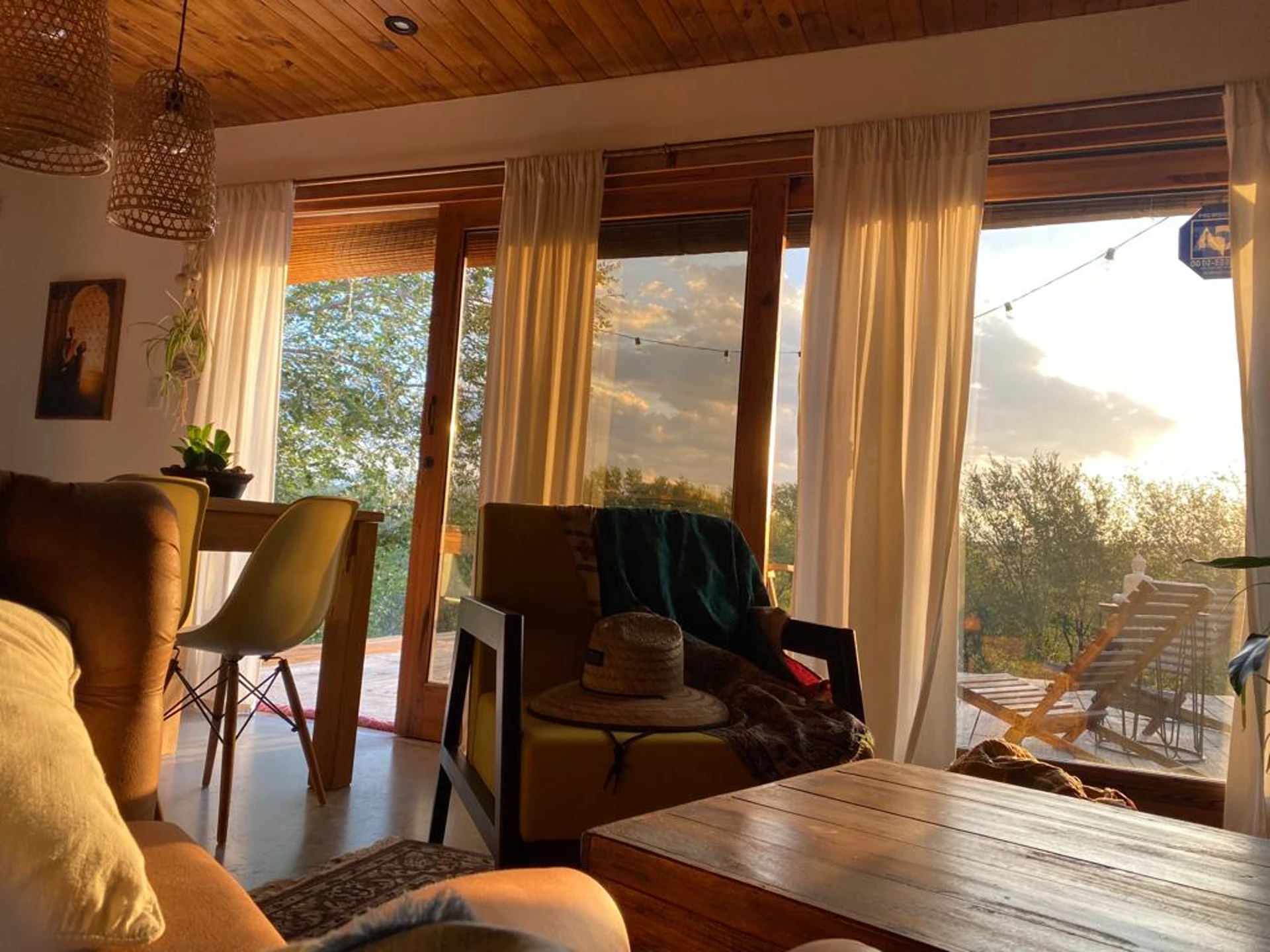 Foto de una casa en Bialet Massé construída con el sistema de Steel Framing por Vértice Construcciones en la que se observa una sala de estar interior con un sector de sillones y muebles de madera con una alfombra y otro sector con una mesa de madera y sillas tipo comedor y dos lámparas colgantes de mimbre; detrás hay un gran ventanal a través del cual se observa un deck de madera con muebles de exterior y una guirnalda de luces