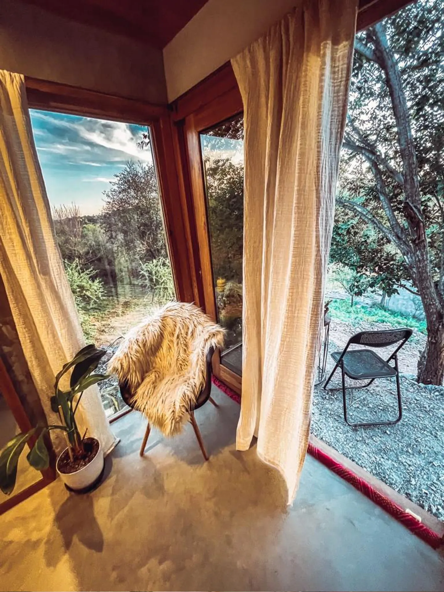 Foto de una casa en Bialet Massé construída con el sistema de Steel Framing por Vértice Construcciones en la que se observa un ángulo interior de la casa con ventanas de piso a techo con aberturas de madera y una silla; a través de las ventanas se observa un paisaje agreste con árboles y arbustos de clima seco en el exterior