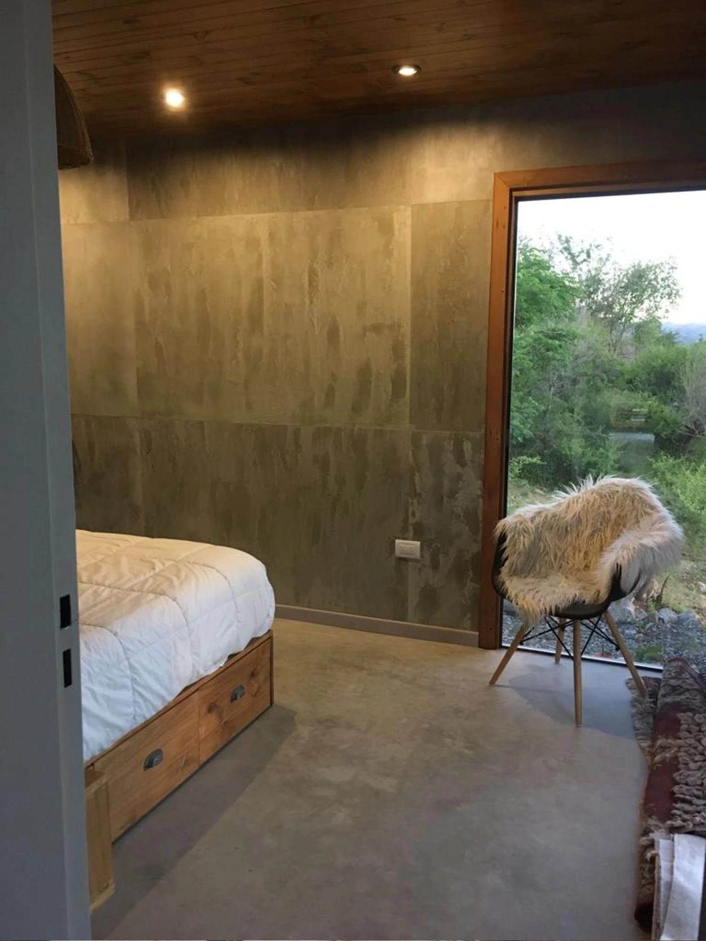 Foto de una casa en Bialet Massé construída con el sistema de Steel Framing por Vértice Construcciones en la que se observa un dormitorio con una cama matrimonial y una silla; el piso es de hormigón alisado color gris plomo, el techo de madera y hay una ventana con abertura de madera a través de la cual se observa un paisaje agreste con árboles y arbustos de clima seco en el exterior