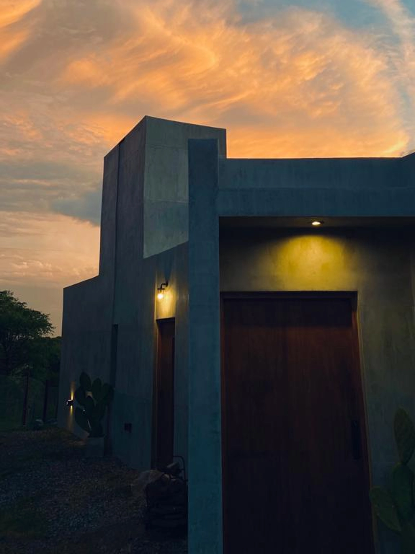 Foto de una casa en Bialet Massé construída con el sistema de Steel Framing por Vértice Construcciones en la que se observa la fachada principal de la casa iluminada artificialmente ya que es de noche, con un acabado símil hormigón visto y una puerta principal de madera lisa, y varias macetas de hormigón con plantas de clima seco como cactus y espinillos