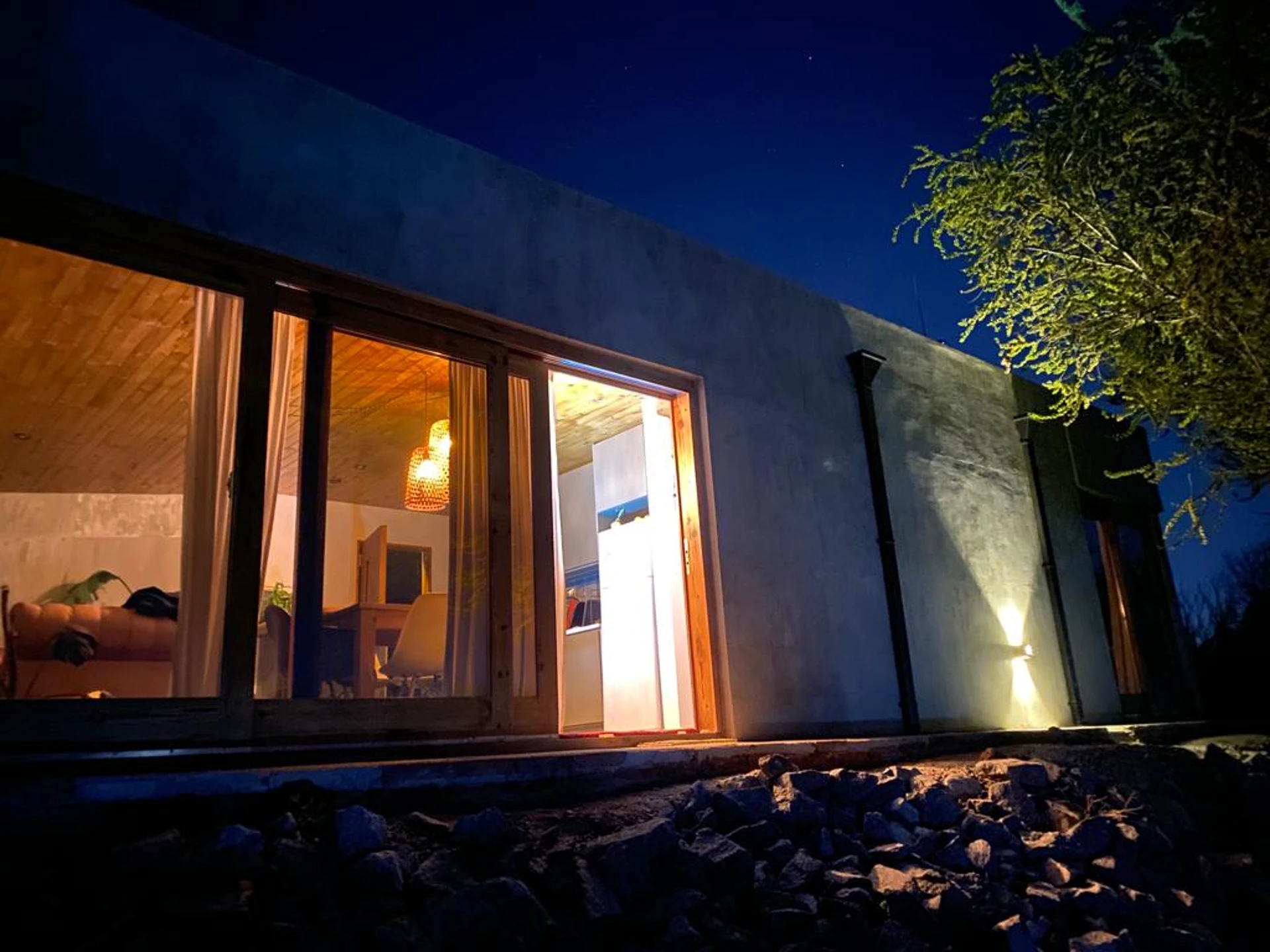 Foto de una casa en Bialet Massé construída con el sistema de Steel Framing por Vértice Construcciones en la que se observa la fachada trasera de la casa iluminada artificialmente ya que es de noche, con un acabado símil hormigón visto y una puerta-ventana con aberturas de madera; se puede observar también en el interior una sala de estar iluminada con un sillón y una mesa de madera con sillas