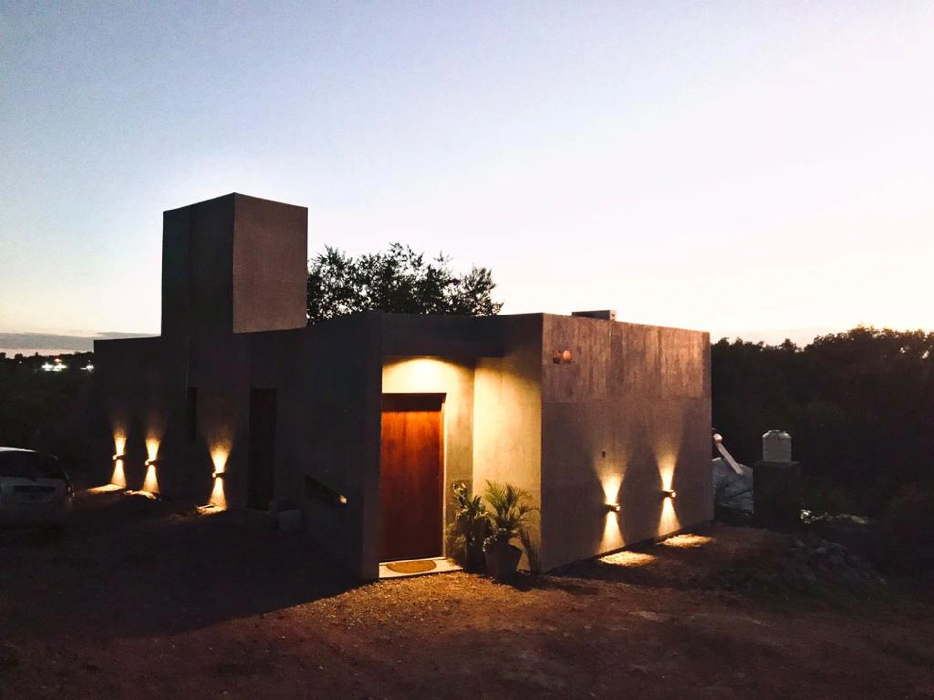 Foto de una casa en Bialet Massé construída con el sistema de Steel Framing por Vértice Construcciones en la que se observa la casa iluminada artificialmente ya que es de noche, con la puerta prinipal de madera con dos macetas y plantas y un acabado símil hormigón visto, y otra puerta secundaria en otra cara de la casa