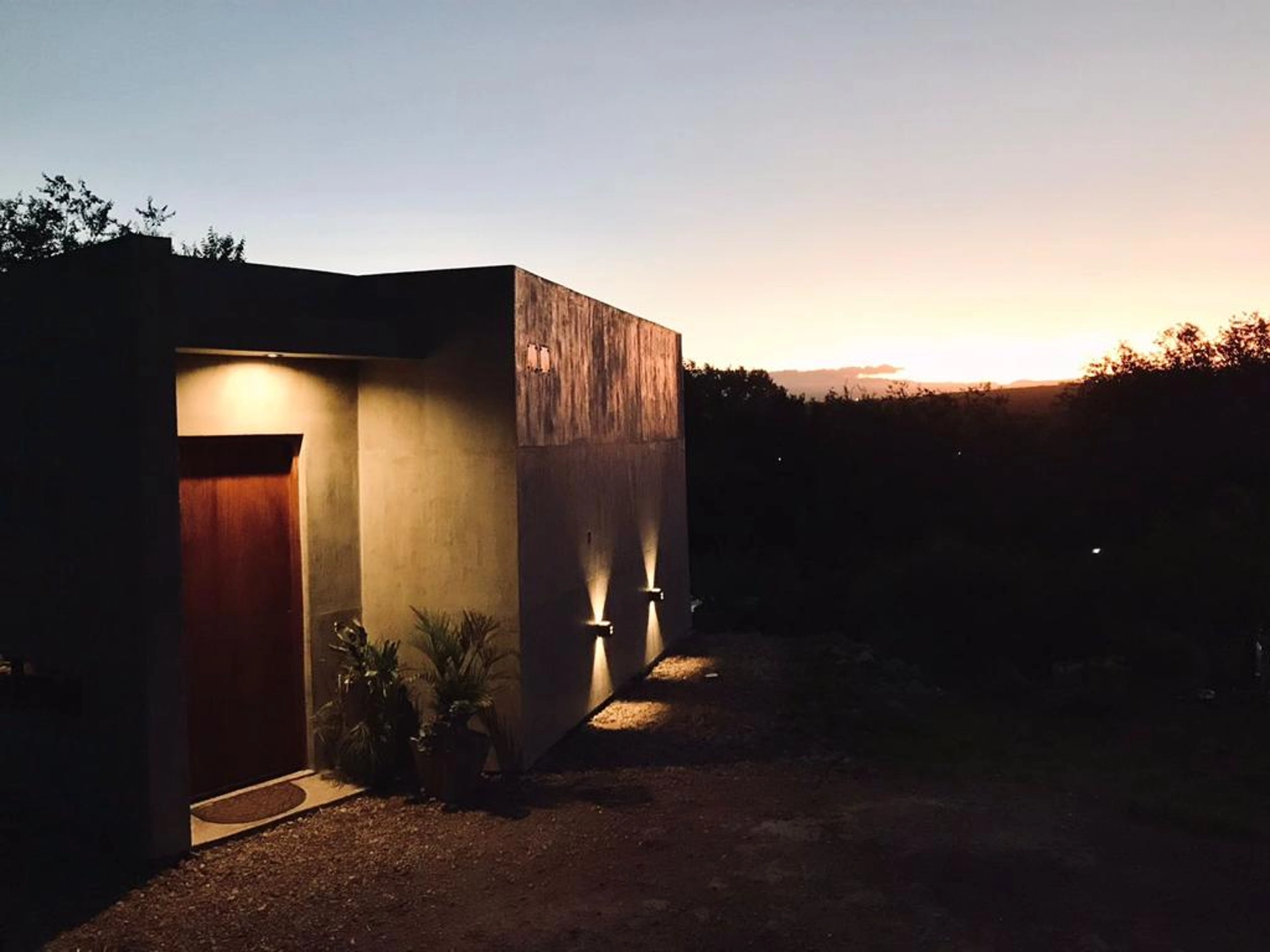 Foto de una casa en Bialet Massé construída con el sistema de Steel Framing por Vértice Construcciones en la que se observa la casa iluminada artificialmente ya que es de noche, con la puerta prinipal de madera con dos macetas y plantas y un acabado símil hormigón visto