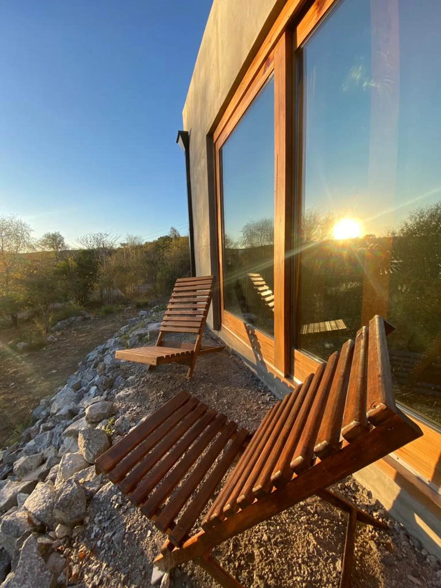 Foto de una casa en Bialet Massé construída con el sistema de Steel Framing por Vértice Construcciones en la que se observa parte de la fachada de la casa con un acabado símil hormigón visto y una puerta-ventana con aberturas de madera y dos sillas de madera en el exterior