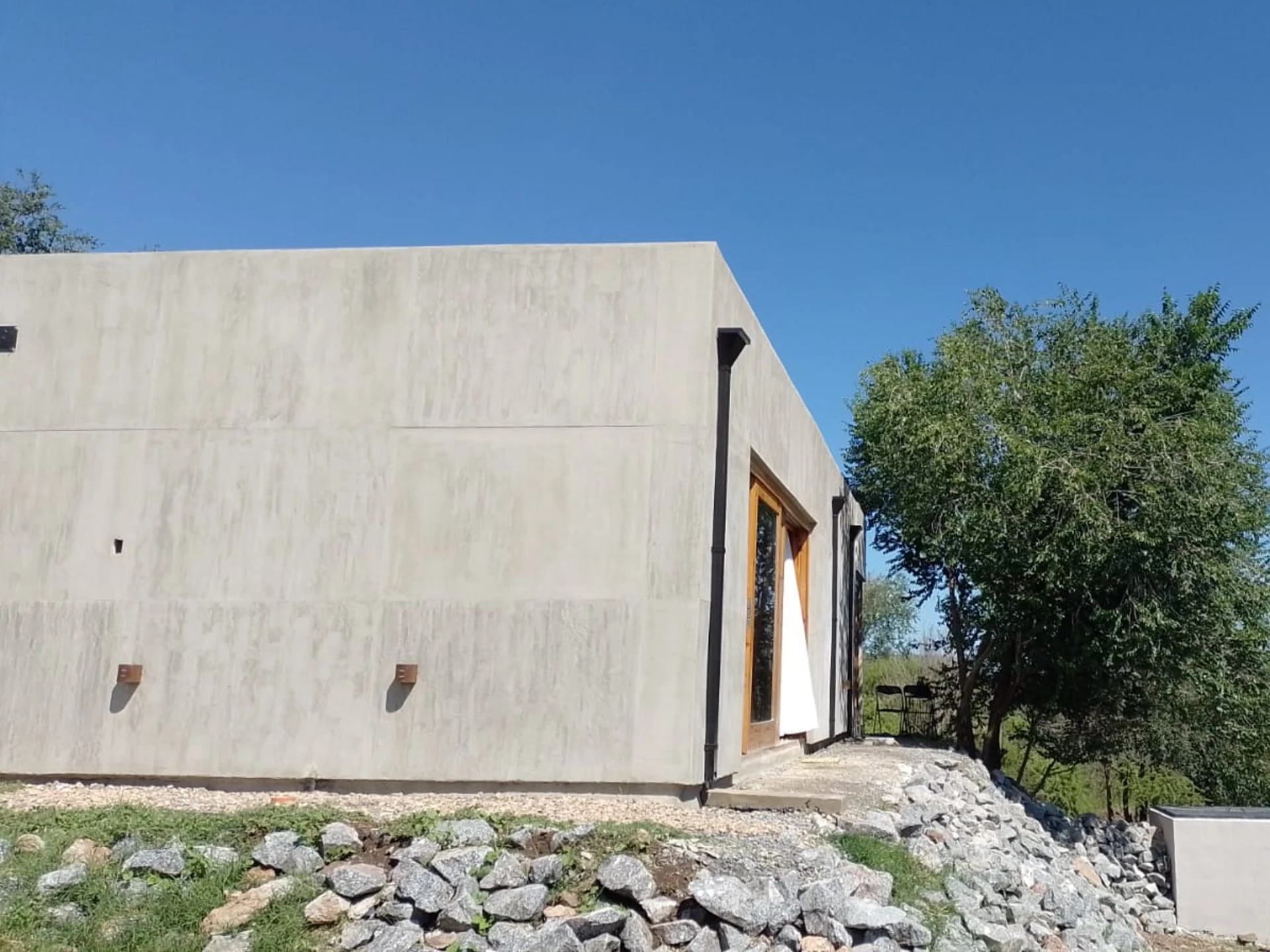 Foto de una casa en Bialet Massé construída con el sistema de Steel Framing por Vértice Construcciones en la que se observa un ángulo de la casa con un acabado símil hormigón visto y una puerta-ventana con aberturas de madera, y un árbol en una esquina de la casa