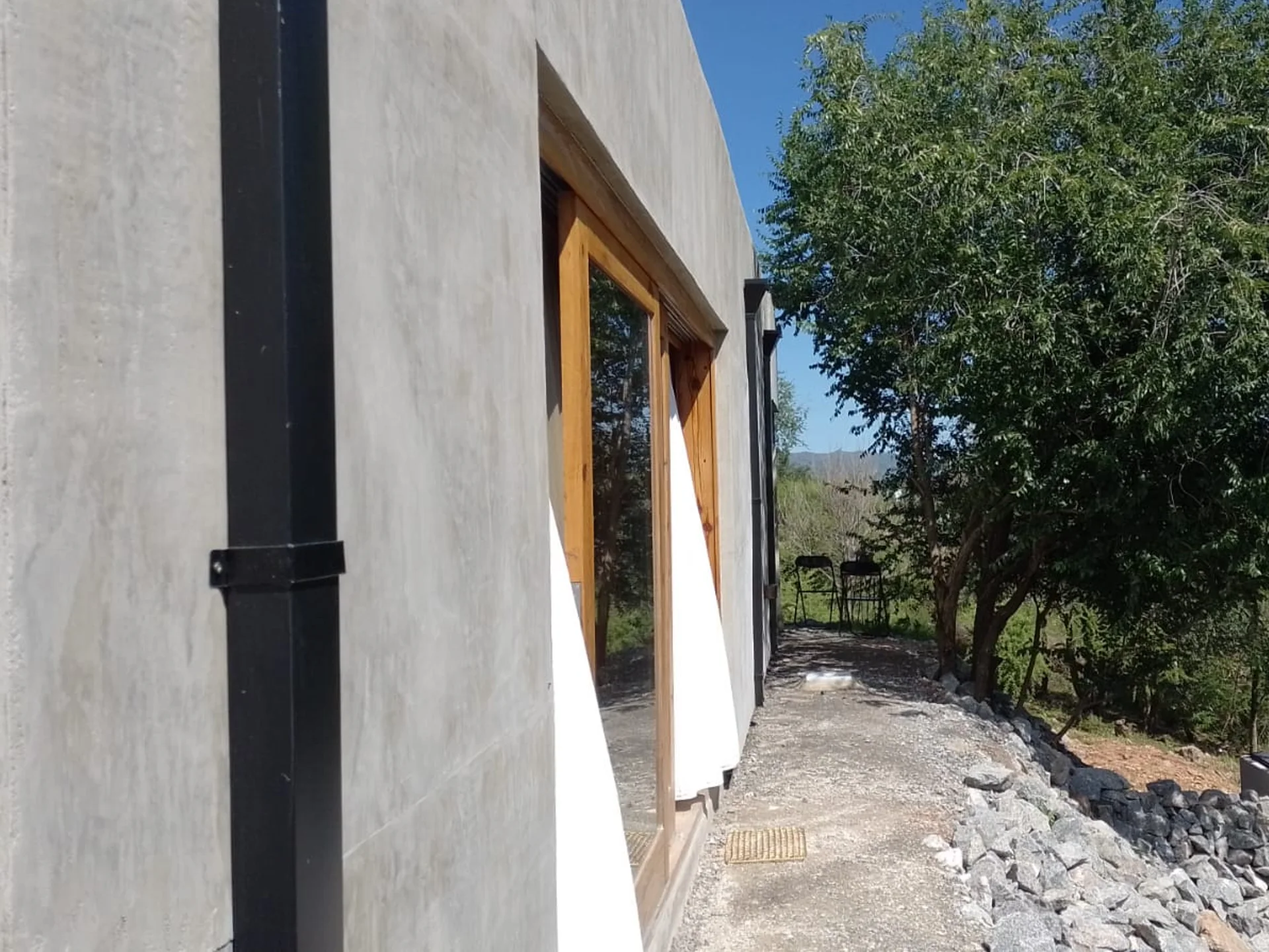 Foto de una casa en Bialet Massé construída con el sistema de Steel Framing por Vértice Construcciones en la que se observa en primer plano parte de la fachada trasera de la casa con un acabado símil hormigón visto y una puerta-ventana con aberturas de madera, y un árbol en una esquina de la casa