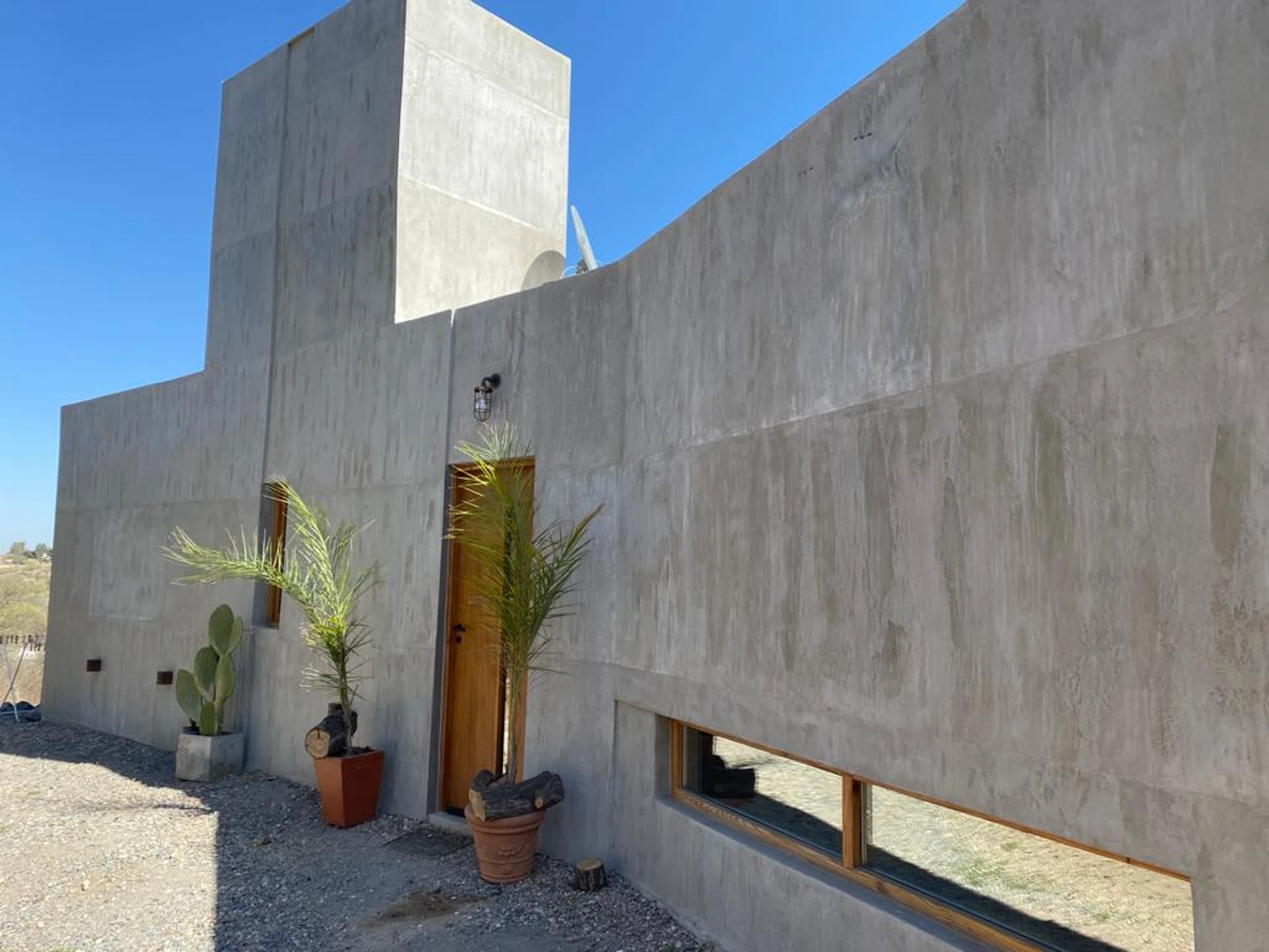 Foto de una casa en Bialet Massé construída con el sistema de Steel Framing por Vértice Construcciones en la que se observa una fachada de la casa con un acabado símil hormigón visto, una puerta de madera con un vidrio vertical estrecho flanqueada por dos macetas con un árbol pequeño cada una, y dos ventanas con aberturas de madera