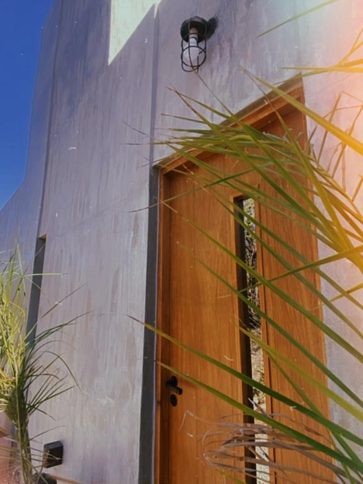 Foto de una casa en Bialet Massé construída con el sistema de Steel Framing por Vértice Construcciones en la que se observa en primer plano una puerta de madera con un vidrio vertical estrecho y parte de una fachada de la casa con un acabado símil hormigón visto